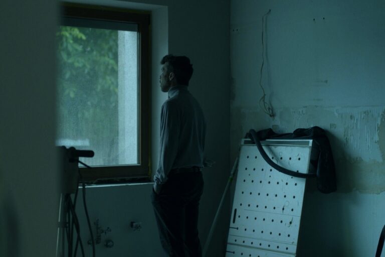 Kadr z filmu "Dzień Ojca". Mężczyzna wygląda przez okno w pomieszczeniu w stanie do remontu, ze skutymi kafelkami i gołymi ścianami z widocznymi kablami.
