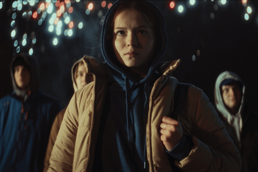 Kadr z filmu "Fiber". Dziewczyna patrzy przed siebie, ma zdeterminowany wyraz twarzy. Za jej plecami stoi kilka osób w podobnym jej wieku.