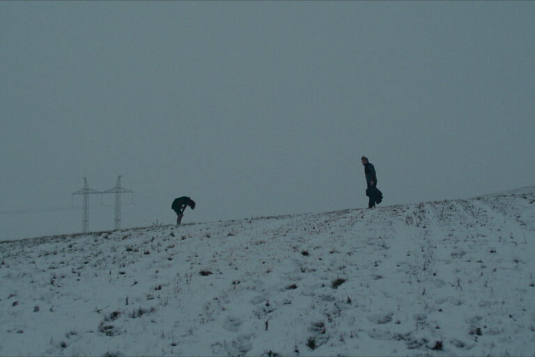 Kadr z filmu "Gone". Dwie postacie na przykrytej śniegiem łące. Jedna zgina się wpół.