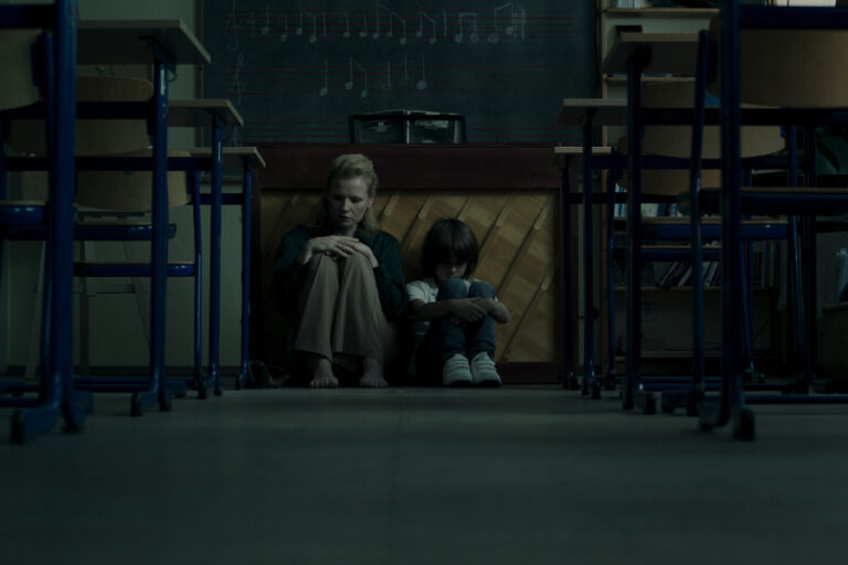 Kadr z filmu "I Gaze at the Sky". Kobieta i dziecko siedzą ze spuszczonymi głowami na podłodze szkolnej sali.