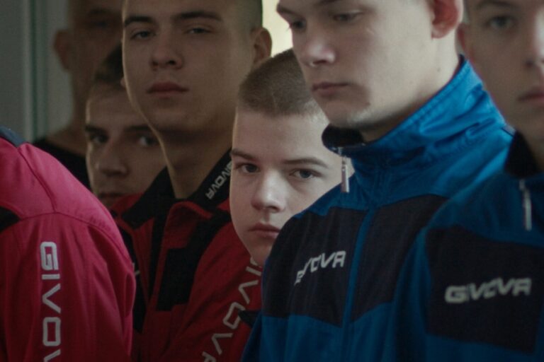 Kadr z filmu "Świeżak". Młodszy chłopak stoi w grupie starszych chłopaków, ma obojętny wyraz twarzy.