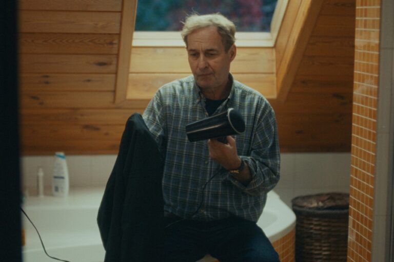 Kadr z filmu "Syn szczęścia". Mężczyzna siedzi na skraju wanny i suszy suszarką marynarkę lub sweter.