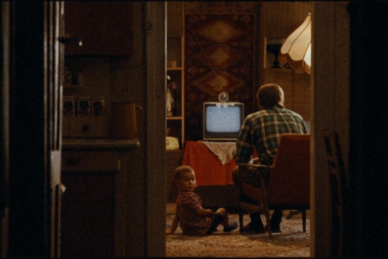 Kadr z filmu "Taniec w narożniku". Mężczyzna siedzi w fotelu plecami do kamery, wpatrzony w stary telewizor. U jego stóp na podłodze siedzi chłopiec, patrzący w stronę kamery.